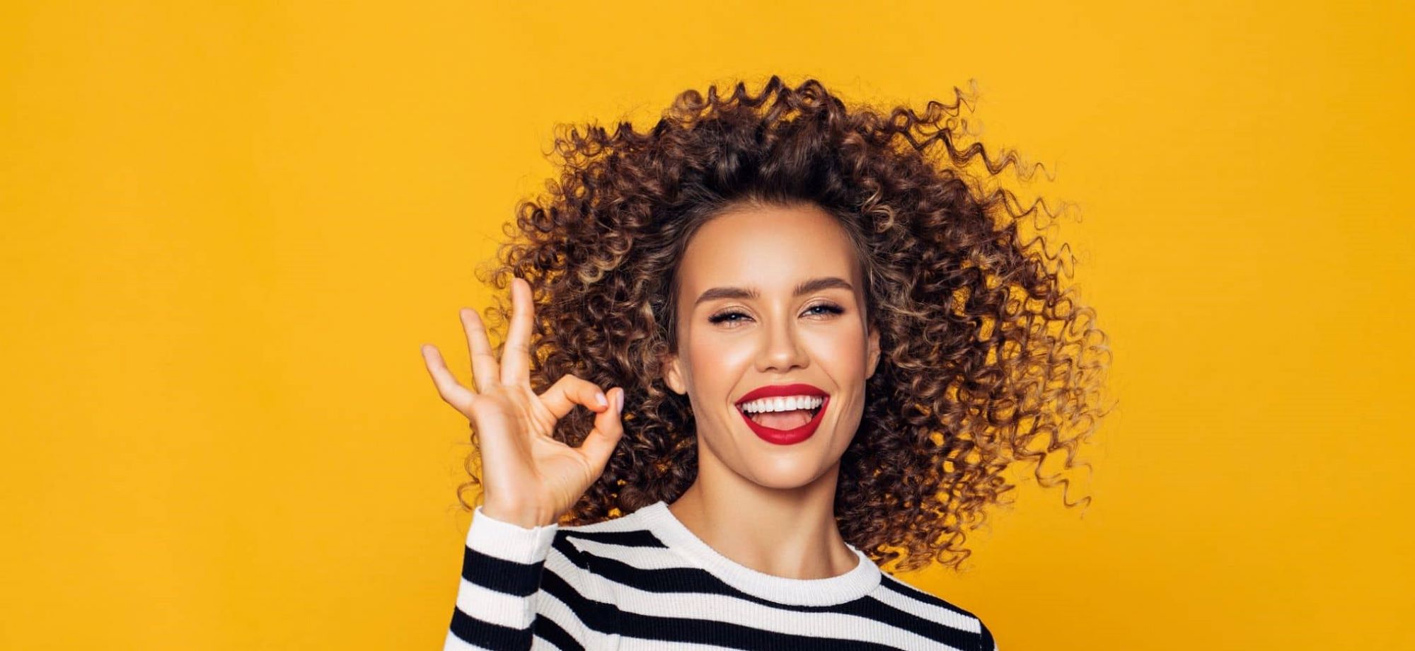 beautiful woman with curly hair and huge smile in front of mustard yellow background